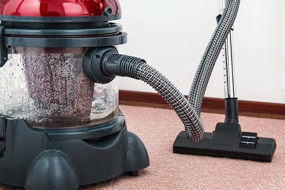 Close-up of a vacuum cleaner with a transparent dust container filled with debris, connected to a flexible hose that is attached to a cleaning head on a pink carpeted floor. The vacuum cleaner has a r