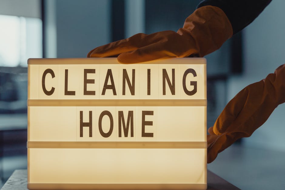 A person wearing orange cleaning gloves is placing a lightbox sign with black letters spelling 'CLEANING HOME' on a table in a modern, well-lit room. The surface of the table appears smooth and clean, and the background shows a blurred interior with natural daylight coming in. This image visually represents domestic surface cleaning and deep cleaning activities performed by professionals like Hounslow Carpet Cleaners, highlighting the importance of proper hygiene and maintenance during end of tenancy cleaning processes in residential spaces.