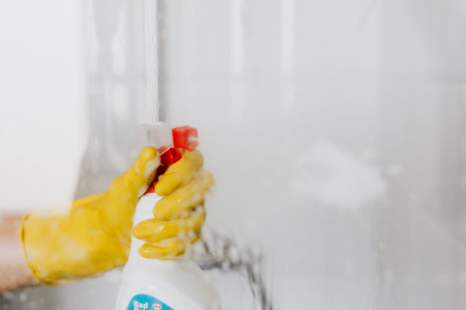 Close-up of a person's hand wearing a yellow rubber glove, holding a spray bottle of cleaning solution. The spray bottle has a white body with a red nozzle and a blue label, and the person's hand is pressing the trigger, releasing a fine mist of cleaning spray. The background features a partly visible white tiled wall and countertop, suggesting a kitchen or bathroom area prepared for surface cleaning as part of domestic or commercial sanitisation efforts. The scene emphasizes thorough cleaning processes that can address common issues during end of tenancy cleaning, as carried out by Hounslow Carpet Cleaners.