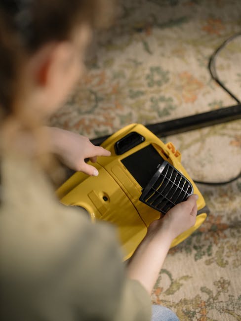 A person holding a yellow portable air mover fan is positioned on a patterned rug, likely during a surface cleaning or drying process. The room has a wooden floor and some visible electrical cords. The individual's hand is near the control panel of the equipment, and the scene appears well-lit with natural or artificial light, emphasizing the cleanliness and readiness for further cleaning or sanitisation. This image is associated with Hounslow Carpet Cleaners, matching the context of domestic or commercial deep cleaning as described in the Hounslow High Street carpet cleaning guide.