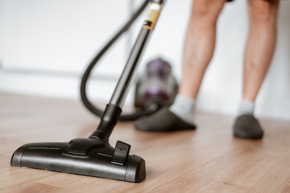 Close-up of a black vacuum cleaner head resting on a wooden floor during a domestic cleaning session, with the blurred figure of a person in shorts and socks operating the appliance. The room has natural lighting, and a purple vacuum canister is visible in the background. The scene highlights surface cleaning and deep cleaning practices typical of residential sanitisation, emphasizing cleanliness and hygiene, and is associated with Hounslow Carpet Cleaners' services promoted on the page 'Lampton Park home cleaning tips for Hounslow residents'.