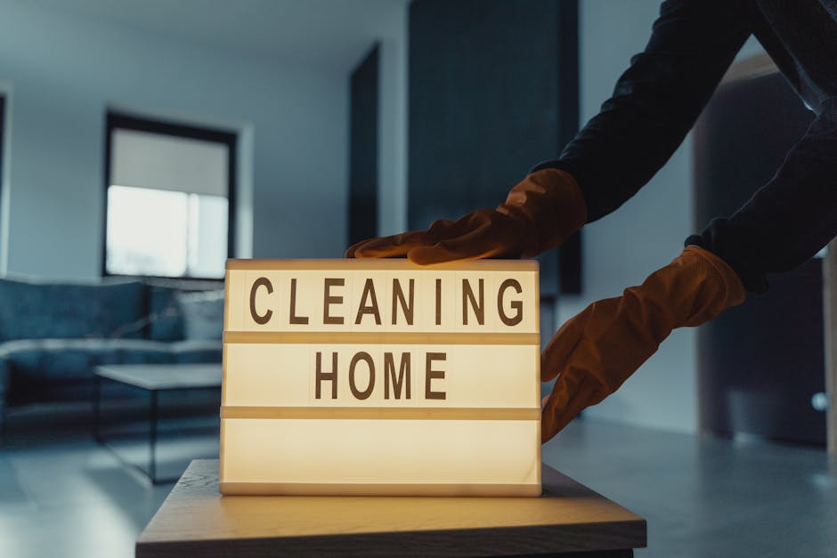 A person wearing orange cleaning gloves is adjusting a small illuminated signboard on a table that displays the words 'CLEANING HOME' in bold black letters. The background shows a modern living room with large windows, a grey sofa, and a coffee table, all appearing tidy and clean. The room is lit naturally, emphasizing the spotless surfaces and well-maintained space, indicative of professional domestic cleaning services provided by Hounslow Carpet Cleaners as part of their surface cleaning and deep cleaning solutions for residents in the Lampton Park area.