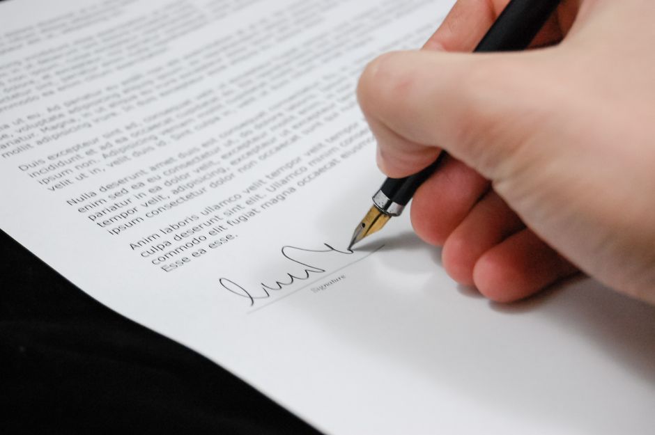 A close-up of a person's hand holding a black fountain pen, signing a document on a white sheet of paper. The paper contains printed text in Latin placeholder format, with the signature being added at