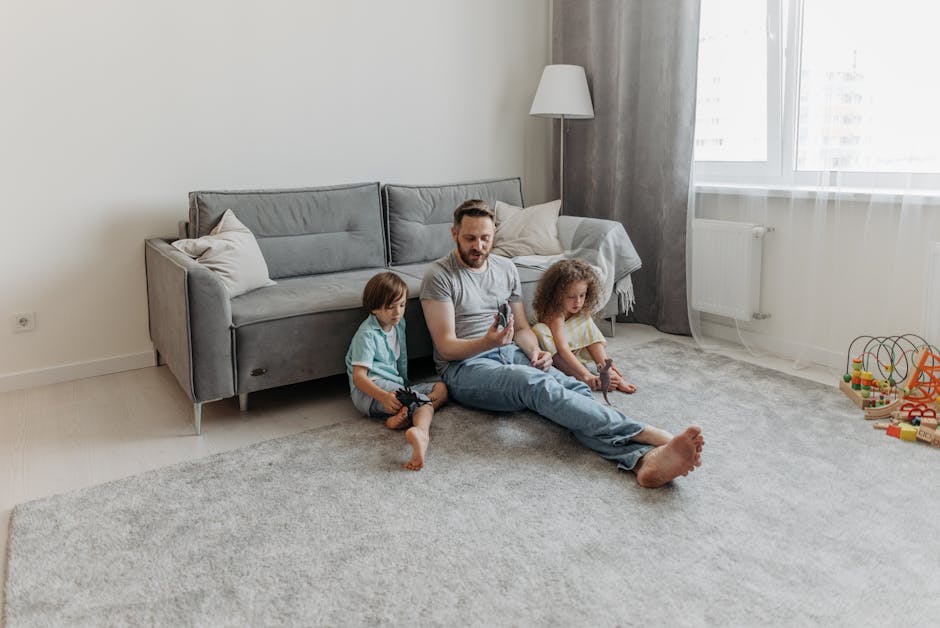 A yellow vacuum cleaner with a black hose attachment is placed on a beige, patterned area rug in a living room. The room has wooden flooring and features a modern black metal and wood TV stand with a