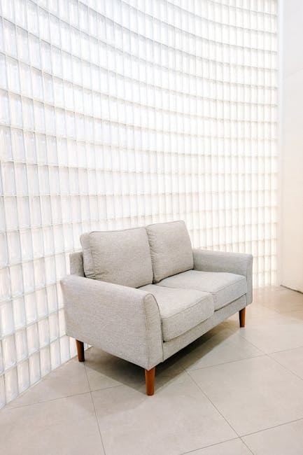 A light grey fabric sofa with wooden legs positioned in a room with beige tiled flooring. The sofa has two large cushions and is placed against a wall featuring a geometric pattern of white, textured, square-shaped panels, creating a modern, clean look. The room is well-lit, emphasizing the pristine and tidy condition of the furniture and surroundings, reflecting professional surface cleaning and upholstery sanitisation by Hounslow Carpet Cleaners as part of their domestic cleaning services for the Treaty Centre area in Hounslow.