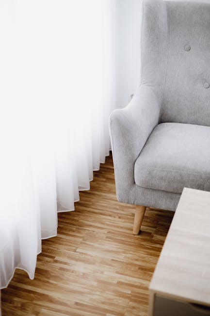 A close-up of a light grey upholstered armchair with a smooth fabric finish, positioned near a set of white sheer curtains that hang to the floor. The armchair has a rounded armrest and a button-tufted backrest, showcasing a clean and well-maintained appearance. The room features a wooden floor with a natural finish and a light-colored wooden side table partially visible in the foreground. Bright, natural light filters through the curtains, illuminating the space evenly. This setting exemplifies a neat and hygienic environment, which Hounslow Carpet Cleaners offers through surface cleaning and domestic cleaning services, ensuring a fresh and sanitized atmosphere.