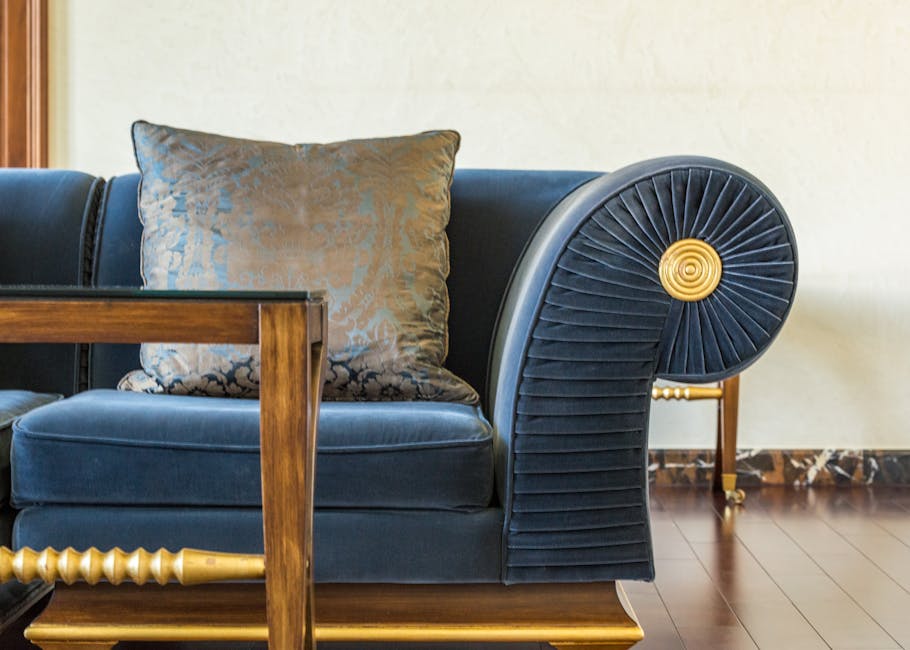 A light grey fabric sofa with wooden legs positioned in a room with beige tiled flooring. The sofa has two large cushions and is placed against a wall featuring a geometric pattern of white, textured, square-shaped panels, creating a modern, clean look. The room is well-lit, emphasizing the pristine and tidy condition of the furniture and surroundings, reflecting professional surface cleaning and upholstery sanitisation by Hounslow Carpet Cleaners as part of their domestic cleaning services for the Treaty Centre area in Hounslow.