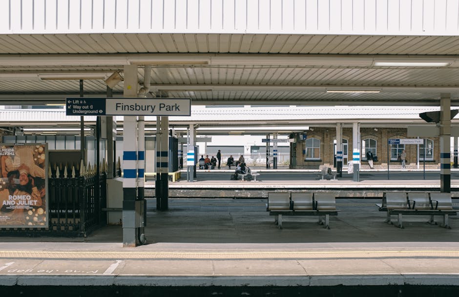 The image depicts a clean, modern train station platform at Finsbury Park, with a covered waiting area supported by metal columns, and several benches for passengers. The platform floor appears smooth and free of debris, with clearly marked spaces and tactile paving near the platform edge. Overhead, a large sign indicates the station name, while smaller signage directs passengers to lifts and underground connections. In the background, commuters are seated on benches or walking along the platform, and a building with large windows and brick walls is visible further down. The scene is well-lit with natural daylight, highlighting the tidy appearance of the station, aligning with the cleanliness standards promoted by Hounslow Carpet Cleaners for surface cleaning and sanitisation of public spaces.