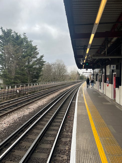 Interior view of a covered walkway near Hounslow West station, featuring smooth concrete flooring with tactile paving strips, illuminated overhead lighting panels, and large glass panels allowing natural light to brighten the space. A person is visible in the distance, standing near an open exit, with greenery and buildings visible outside. The area appears clean and well-maintained, highlighting the importance of surface cleaning and hygiene in public transport environments, which Hounslow Carpet Cleaners can support through professional deep cleaning and sanitisation services.