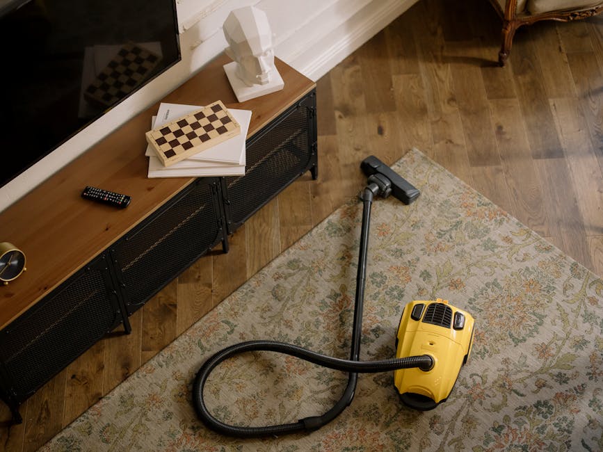 A professional in full protective gear, including a white suit, face mask, and gloves, is performing deep cleaning on a beige carpet in a modern living room. The worker holds a vacuum hose connected to a steam or extraction cleaning device, with visible steam or cleaning mist emanating onto the carpet surface. The room features a light-colored wooden bookshelf with decorative items, a large mirror propped against the wall, and a potted plant. Natural light illuminates the space, highlighting the clean, well-maintained appearance of the carpet and surroundings. This scene exemplifies thorough surface cleaning and sanitisation efforts in a domestic setting, reflecting the services offered by Hounslow Carpet Cleaners.